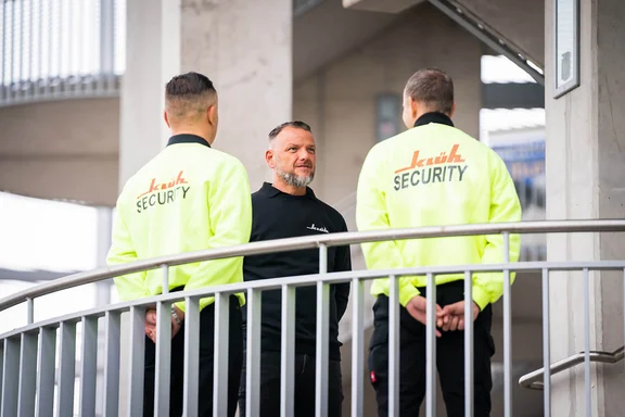 zwei Klüh Security Angestelle in Arbeitskleidung in Warnfarbe Geld sowie einer Schwarzgekleidet stehen zueinander und schauen sich an