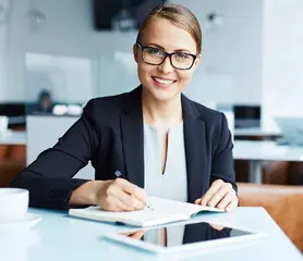 Eine weibliche Person sitzt schreibend an einem Schreibtisch und lächelt.