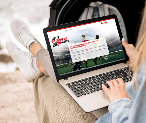 eine Frau hat auf ihren schoß ein Notebook und auf dem Bildschirm ist ein Bild von der Aktion "Tipp-Champion 2024" zu sehen