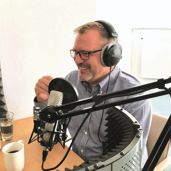Der Klüh Catering Geschäftsführer Thorsten Greth sitzt mit Kopfhörern vor einem Podcast-Mikrofon. Vor ihm stehtein Tisch mit einer Kaffeetasse und im Hintergrund ist ein Fenster zusehen.