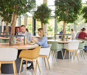 Ein Bild in einer Mensa mit runden Tischen, an denen einige Menschen sitzen und Tellergerichte essen. In der Mitte jedes Tisches befindet sich ein kleiner Baum