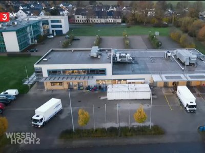 Das Bild zeigt ein Logistikzentrum von oben. Vor dem Zentrum sind 3 LKWs, wovon einer grade wegfährt. 