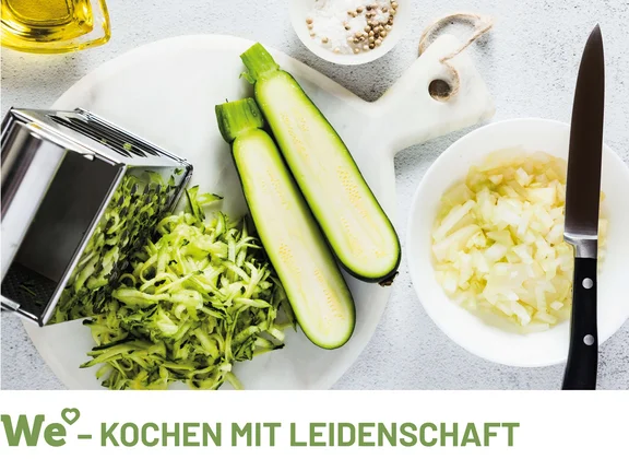 ein Schneidebrett auf dem eine halbierte Zucchini, eine Reibe mit geriebenem Zucchini sowie zwei Schalen mit jeweils geschnittenen Zwiebeln und Salz & Pfeffer und eine kleine Ölflasche mit Öl und dem Untertitel: " We (Herz) - Kochen mit Leidenschaft "