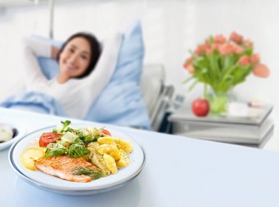 eine Frau liegt im Krankenhaus Bett und schaut auf ein Teller Gericht vor ihr und lächelt dabei