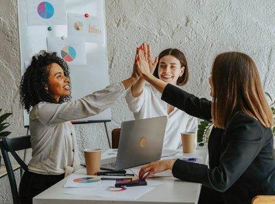 drei Frauen an einem Schreibtisch während eines Meetings lächeln und schlagen zu dritt ein