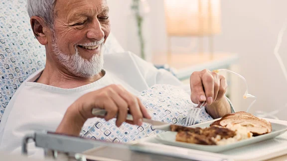 Ein bild von einem Mann im Krankenhausbett und vor sich sein frühstück auf ein Tablett mit einem Glas Orangensaft neben ihm eine Krankenschwester mit einem Klemmbrett und Kugelschreiber