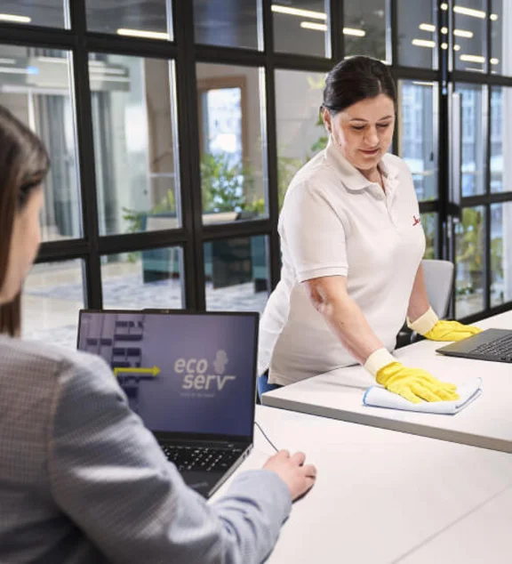 eine Büroangestellte sitzt vor ihrem Notebook an dem Tisch und eine Klüh Reinigungskraft wischt den Tisch mit einem Tuch