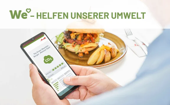 Ein Bild von einem Menschen der sein Handy bedient, auf dem Bildschirm ist die Klüh Catering App geöffnet, im Hintergrund ist ein Burger mit Kartoffelecken auf einem Teller mit Besteck und einer Klüh Serviette. Das Bild hat die Überschrift : WE love - Helfen unserer Umwelt