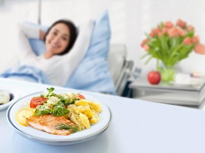 [Translate to Englisch:] Weibliche Person liegt im Krankenbett und sieht lächelnd auf ihr Essen.