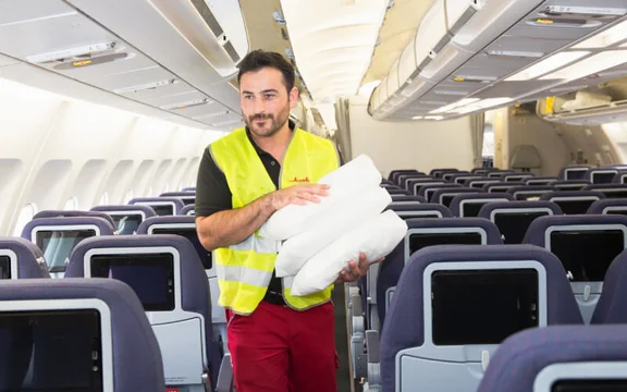 lächelnder Klüh Angesteller in einem leeren Flugzeug mit 3 Kissen in seinen Armen