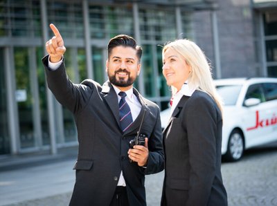 im Hintergrund ein Klüh Multiservices Firmenwagen und davor stehen zwei Klüh Security Angestellten