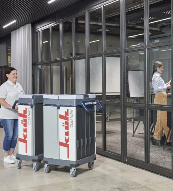 Ein Bild von einer Klüh Cleaning Reinigungskraft meinem Klüh Cleaning Reinigungswagen im Flur eines Bürogebäudes, im Hintergrund ist eine Frau in einem Besprechungsraum mit einer Glaswand 