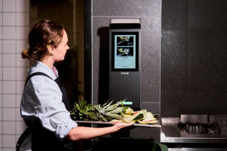 Eine Köchin steht vor dem KI-gestützten Food Waste Gerät Orbisk. Sie hält ein Tablet mit Obst und Gemüse Abfällen in ihren Händen