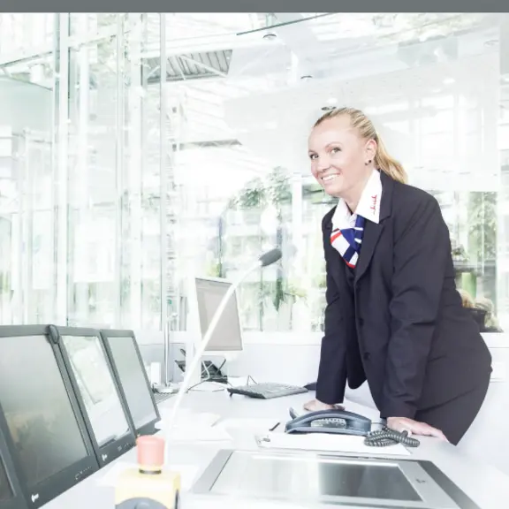 Eine weibliche Servicekraft steht lächelnd an einem Arbeitsplatz mit mehreren Bildschirmen.