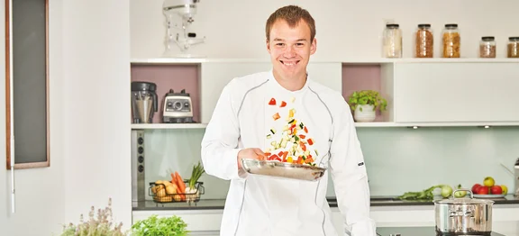 Ein lächelnder Koch in Arbeitskleidung steht in der Küche und schwingt eine Pfanne mit Gemüse drin