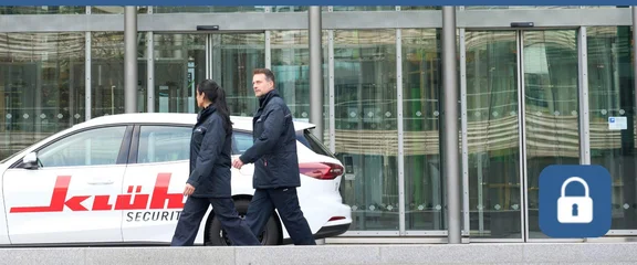 Ein Bild von einem Mann und einer Frau in Klüh Security Arbeitskleidung laufen einem Gebäude mit einer Glasfassade vorbei, vor dem Gebäude parkt ein Klüh Security Firmenwagen, am rechten Rand des Bildes ist ein blaues Icon mit weißen Schlüsselschloß