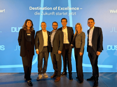 Ein Gruppenbild von 6 Personen in Business Outfits, im Hintergrund ist eine blaue Wand mit dem Schriftzug "Destination of Excellence - die Zukunft startet jetzt"