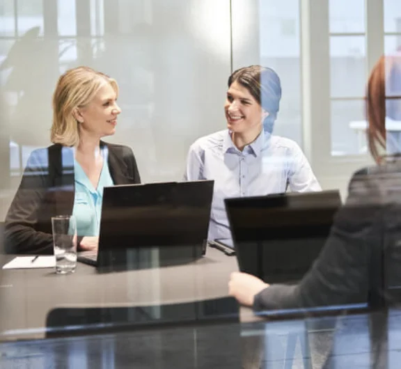 drei Frauen in einem Büromeeting mit ihren Notebooks vor sich auf dem Tisch, die sich lächelnd anschauen 