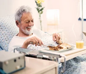 Ein Bild von einem Mann der lächelnd in einem Krankhausbett liegt, vor ihm ein Tablett mit einem Mittagessen und Glas Orangensaft
