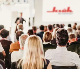 Ein Bild von vielen Menschen die sitzen und auf Bühne schauen, auf der Bühne ist auf der Leinwand das Klüh Multiservices Logo und ein Mann steht davor und redet in richtung der Menschen