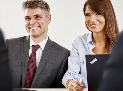 Ein Mann und eine Frau sitzen nebeneinander und sind im Business stil gekleidet, lächeln und schauen in die selbe richtung