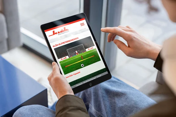 eine Person hat ein Tablet in der Hand und dem Bildschirm ist das Klüh-Elfmeterschiessen zu sehen
