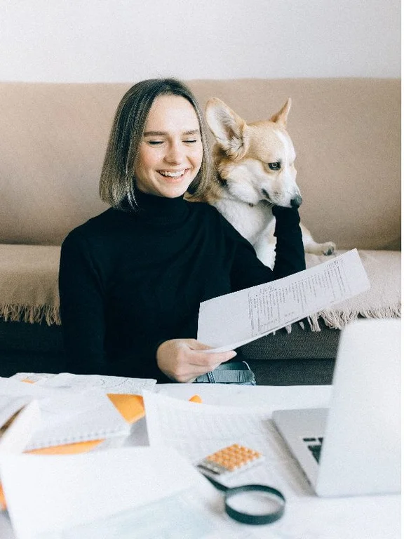 eine lächelnde Frau am Tisch, welcher voller Arbeitsmaterialien und 1 Notebook ist, in einer Hand ist ein Papier und mit der anderen Hand streichelt sie ihren Hund, welcher hinter ihr sitzt