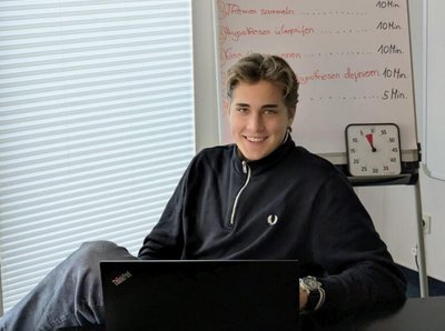 Ein Bild von einem jungen lächelnden Mann während er am Tisch mit seinem Notebook sitzt im Büro