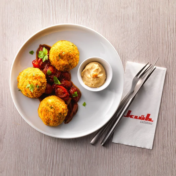 Ein Bild einem Tellergericht auf einem Tisch mit Besteck und einer Klüh Catering Serviette, auf dem Teller sind knusprige Klöße sowie ein Cherrytomatensalat und eine kleine Schale mit einem cremigen Dip