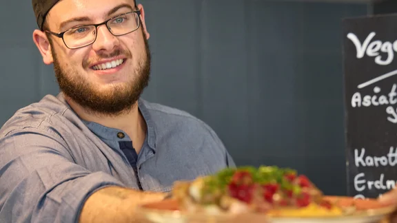 Ein Koch übergibt ein Tellergericht, im Hintergrund ist eine Wand mit dunkelgrauen Fließen und ein Kreidetafel mit der Aufschrift " Vegetarisch Asiatisch gebackene Spitzkohl, Karottenpolenta, Erdnusssauce