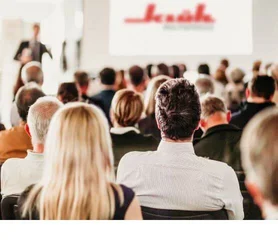 Ein Bild von einem Vortrag von Klüh Multiservices, man sieht viele sitzende Menschen die alle auf die Bühne zum redner schauen