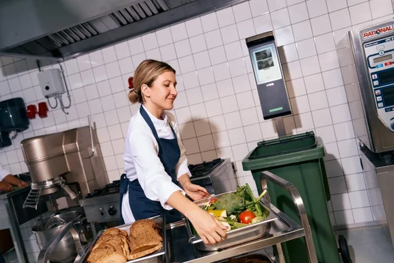 Ein Bild von einer Köchin die lächeln ein Gastro Tablett voller Gemüse und Obst reste anhebt. Im Hintergrund ist das Orbisk Food Waste KI-Gerät