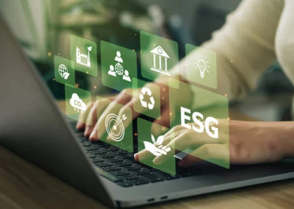 Ein Bild von einer Person die am Schreibtisch sitzt und am Notebook arbeitet, auf dem Bild kommen aus der Tastatur grüne Icons heraus wie: ESG Icon, recycle Icon, Co2 Wolke Icon, Glühbirne Icon und weitere