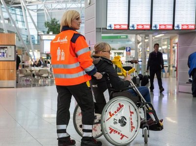 Klüh Multiservices Angestellte in Warnweste schiebt ein Rollstuhl auf dem eine gehbeeinträchtige Frau sitzt durch den Flughafen