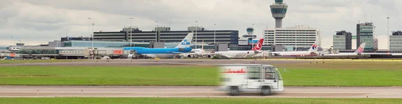 Ein Bild von einem Flughafen Abflug Gelände mit einigen Flugzeugen im Hintergrund und ein Klüh Flughafen Auto, welches die Straße entlang fährt