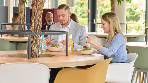 Ein Bild in einer Mensa mit Runden Tischen, wo aus der Tischmitte ein kleiner Baum eingesetzt wurde, an den Tischen sitzen einigen Menschen und essen Tellergerichte. Am Rand des Bildes ist das Klüh Catering Icon