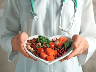 eine Person im Arztkittel und Stethoskop um den Hals hält ein Herzformigen Teller in den Händen, auf dem Teller sind Zutaten wie: Lachs, Brokkoli, Cashews, Walnüsse, Bohnen, Tomate und Knoblauch 