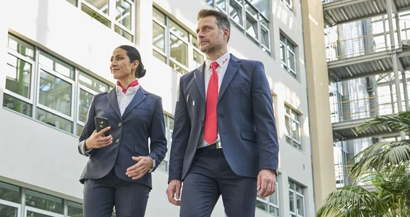 Ein Mann und eine Frau im Anzug mit weißen Hemd mit dem Klüh Logo am Kragen in einem Gebäude