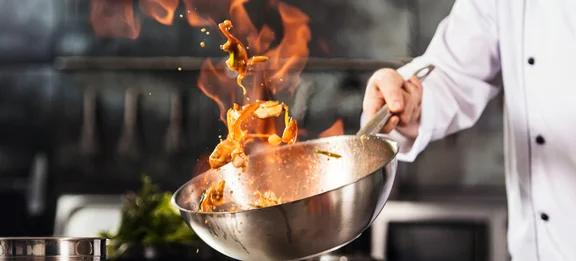 Ein Koch schwenkt ein Wok mit Zutaten die in flammen sind in einer Küche