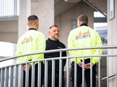 Ein Bild von drei Männdern die sich gegenüberstehen, zwei Männer haben Arbeitskeidung von Klüh Security an in der Farbe gelb und ein Mann trägt ein schwarzes Klüh Security Oberteil