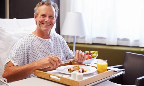 Ein Bild von einem lächelnden Mann in seinem Krankenhausbett hat vor sich sein Mittagsessen auf ein Tablett mit einem Glas Orangensaft