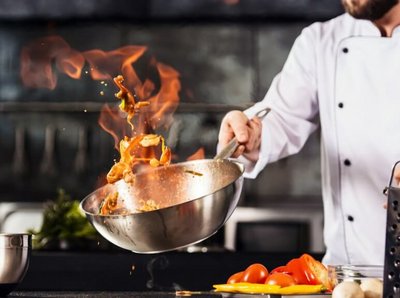 ein Koch schwingt ein Wok mit Zutaten, welche flammen bedeckt sind in einer Küche
