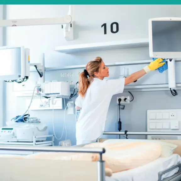 Eine weibliche Reinigungskraft wischt in einem Krankenhauszimmer Oberflächen ab.