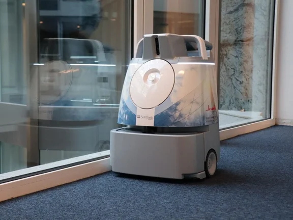 Ein Bild von einem Reinigungsroboter mit einem Klüh Cleaning Aufkeber in einem Flur mit blauen Teppich Boden