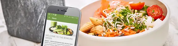 Auf dem Bild ist eine gesunde Bowl mit Salat und ein Smartphone zu sehen, auf dem eine Klüh App in Anwendung ist.