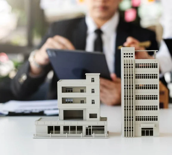 Ein Bild von einem Mann im Anzug der am Schreibtisch sitzt und ein Tablet in der Handy hält, vor ihm auf dem Tisch sind 2 Miniatur Gebäude