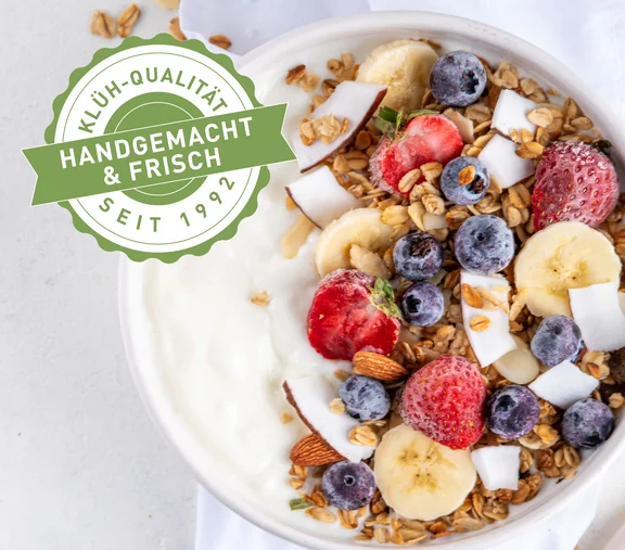 Ein Bild von einer Frühstücksbowl mit Joghurt mit Beeren, Bananenscheiben, Haferflocken und Nüssen und ein Klüh-Qualität Handgemacht & Frisch seit 1992 Siegel