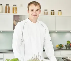 Ein Koch steht an einem Tisch mit Zutaten und lächelt.