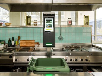 Ein Bild von einer Betriebsküche. Im Vordergrund steht eine grüne Mülltonne und dahinter das KI-gestütze Food Waste Gerät "Orbisk"