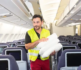 Ein Mann in Warnweste mit Handtüchern auf dem Arm läuft durch einen Flugzeuggang. Links und rechts befinden sich Flugzeugsitze.